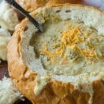 Broccoli Cheese Soup in a round bread bowl topped with shredded cheddar cheese.