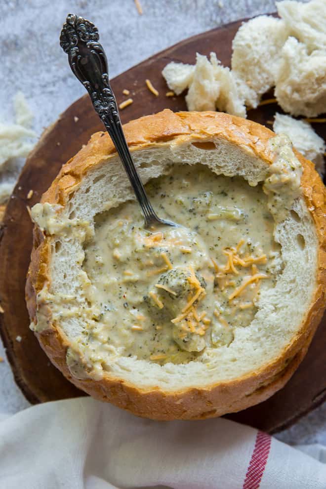 A bread bowl full of Broccoli Cheese Soup with torn pieces of bread next to it.