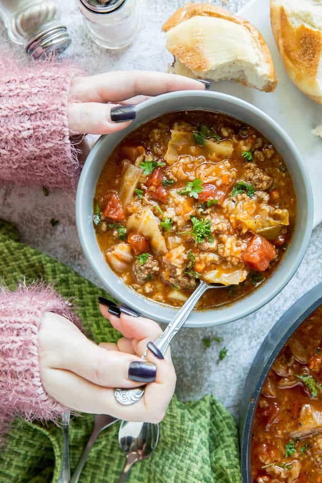 Hands hold the side of the bowl and a spoon is inserted into a bowl of the soup.