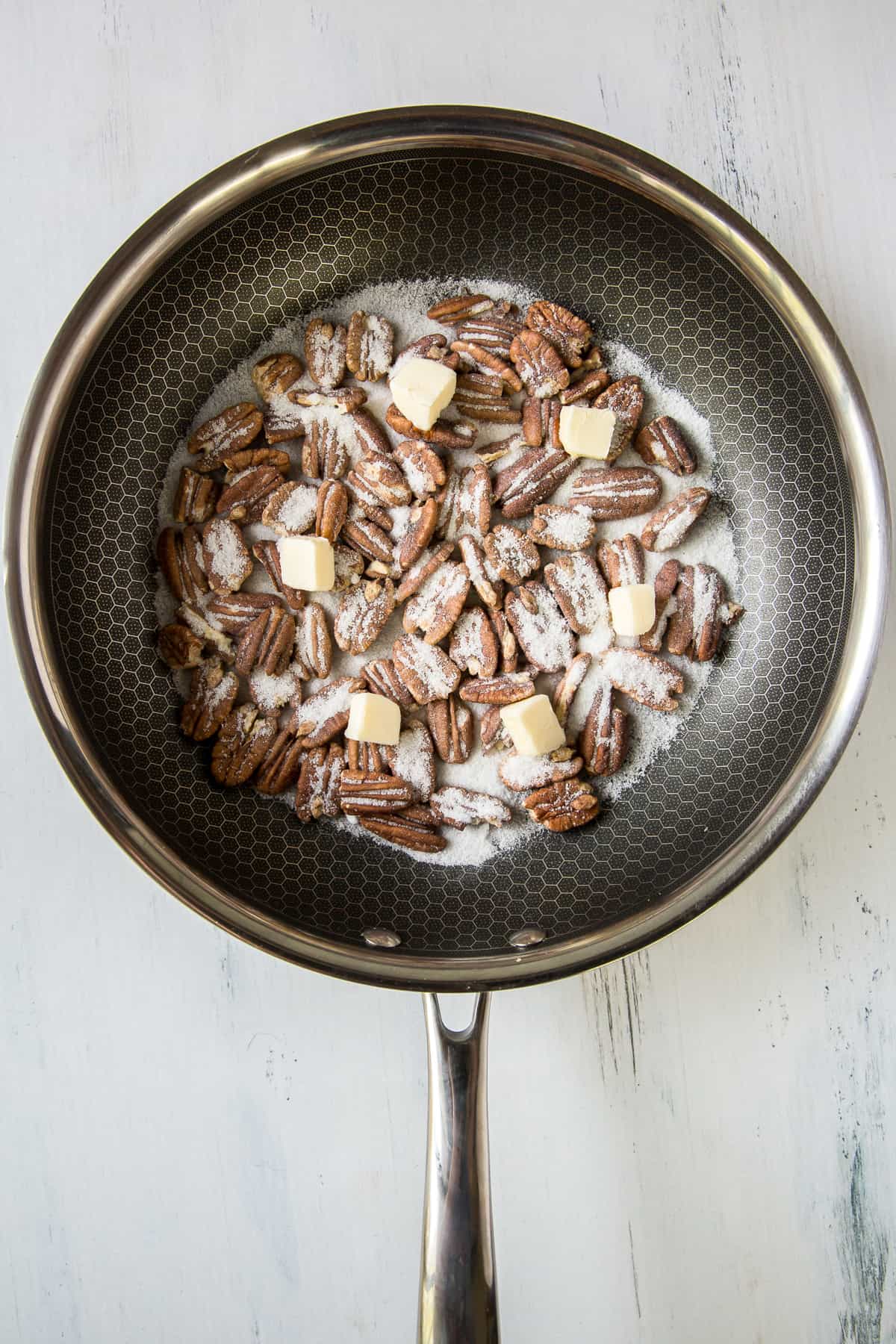 A top down shot of pecans, sugar, and pieces of butter in a skillet.