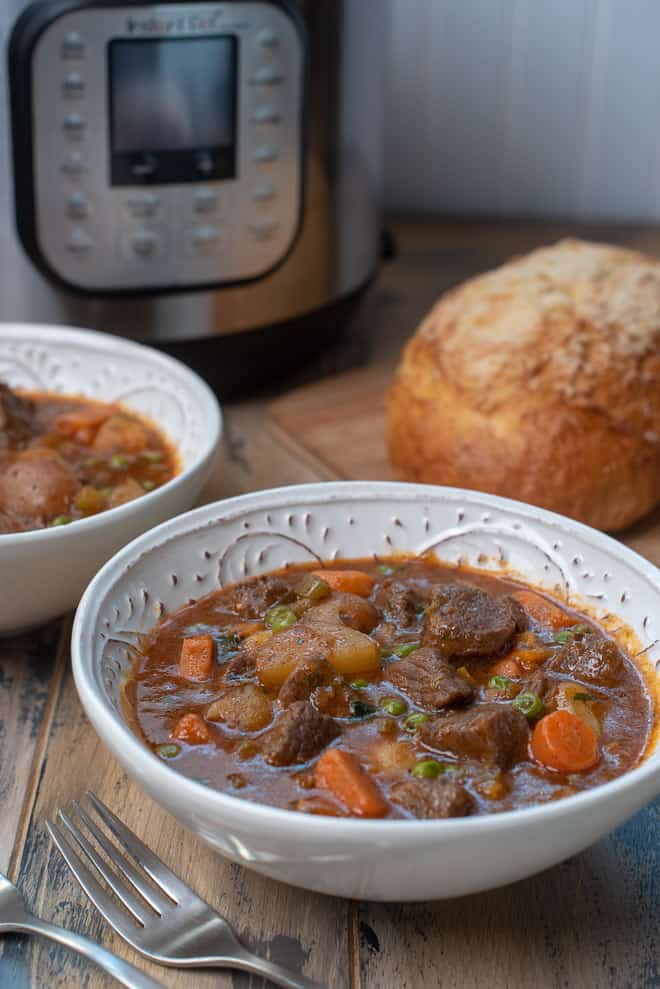 White serving bowls of beef stew with an Instant Pot and French bread in the background.