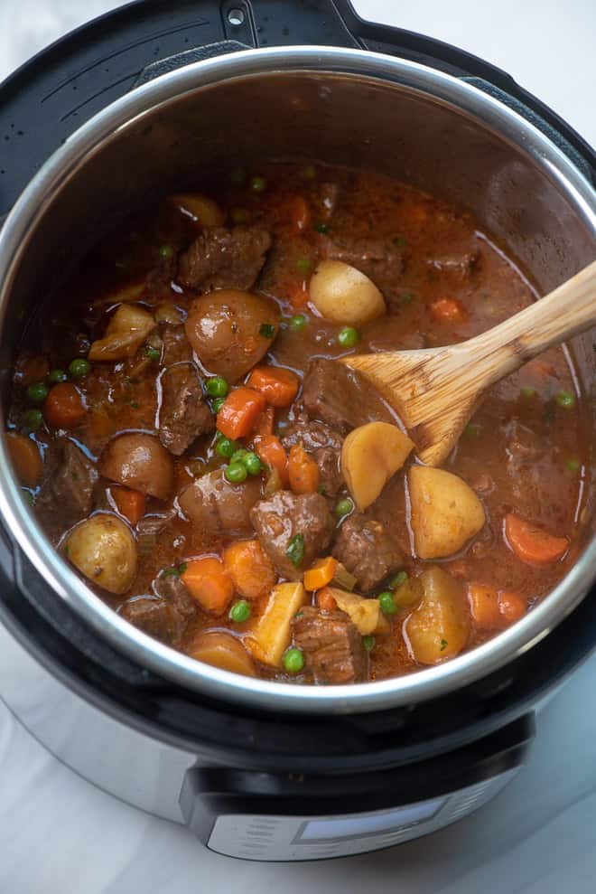 A wooden spoon stirs peas, parsley, and cornstarch slurry into the beef stew in the Instant Pot.