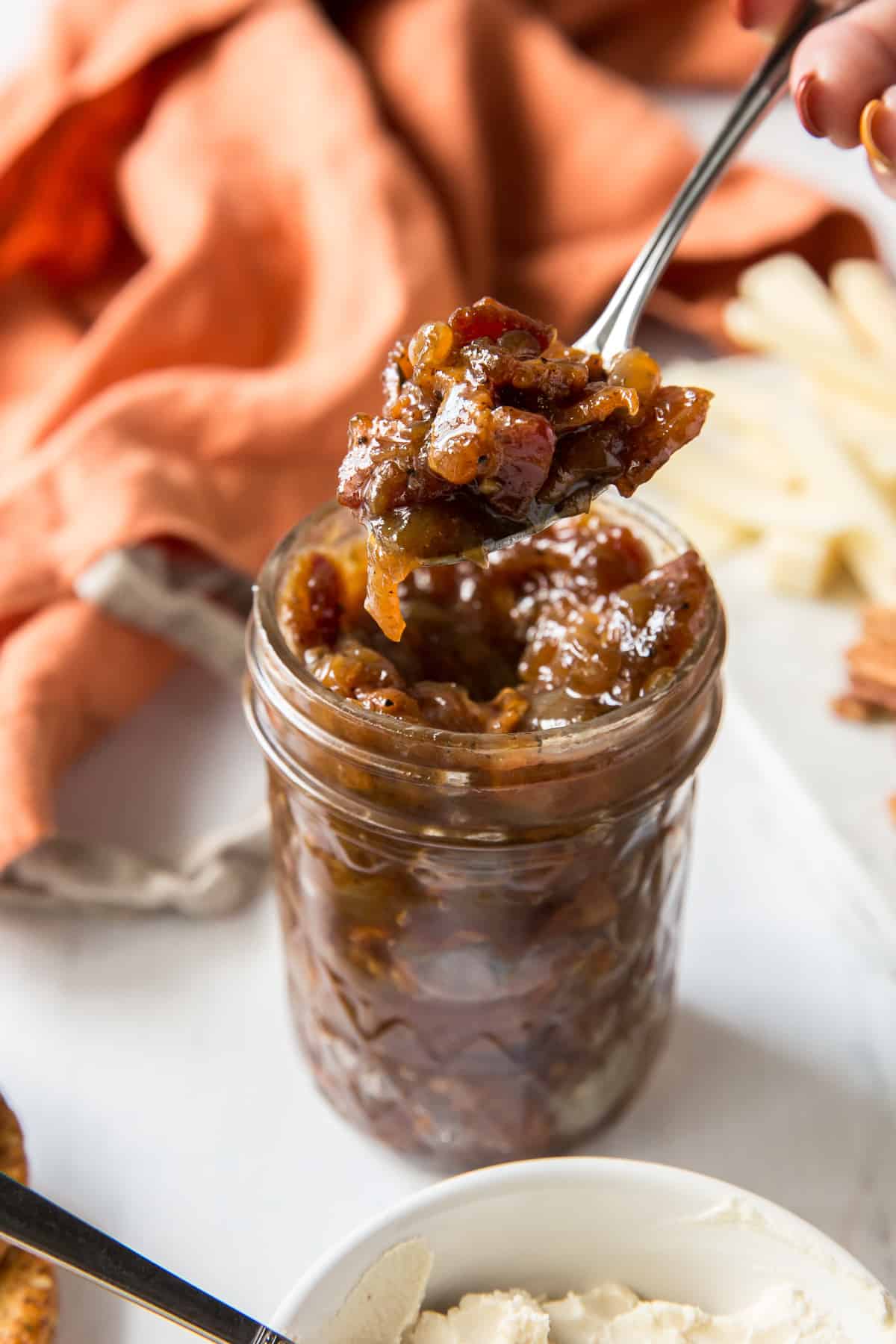 A hand holding a spoon that is hovering over a mason jar filled with bacon jam.