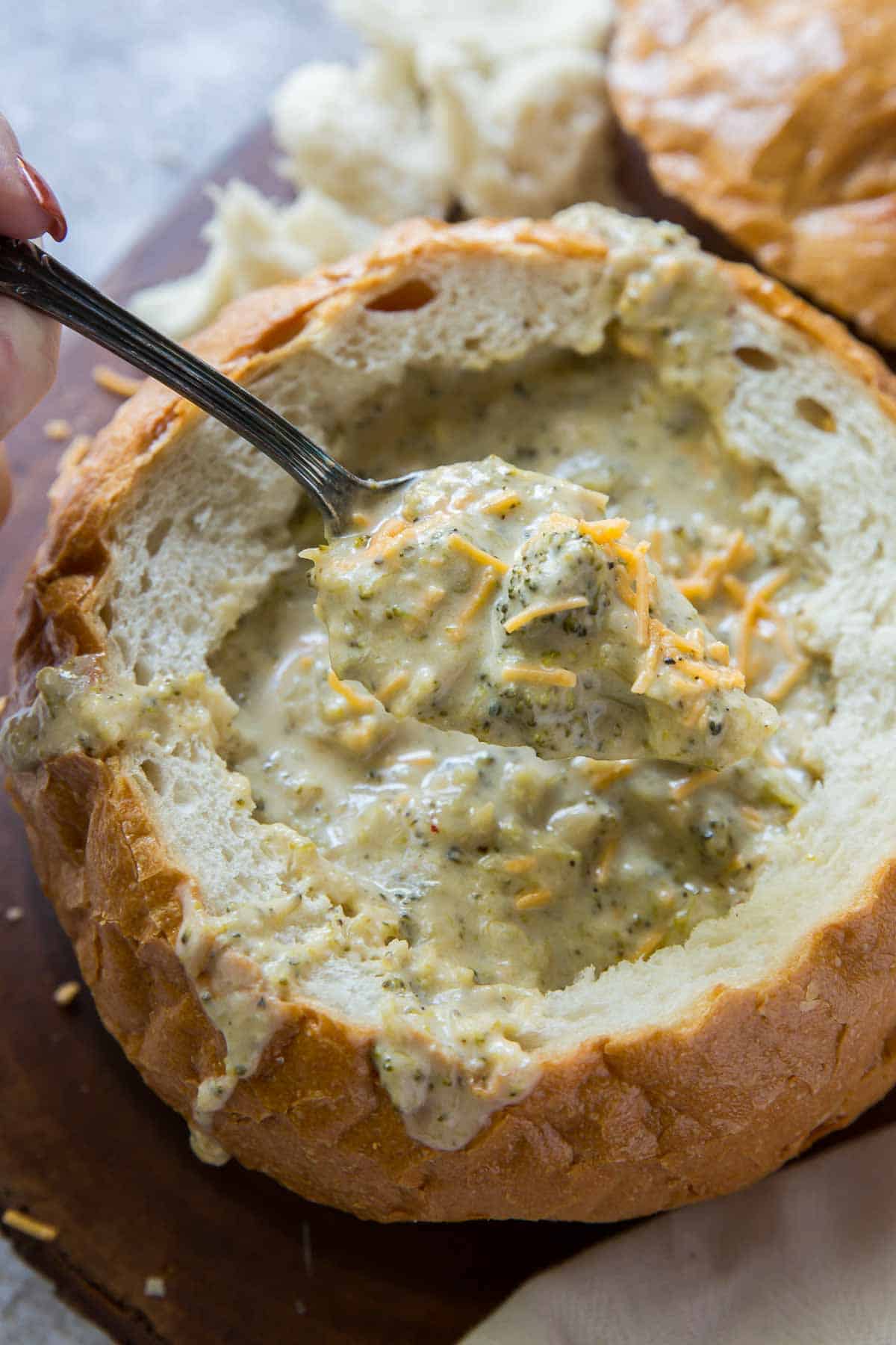 A spoon scooping broccoli cheese soup from a bread bowl.