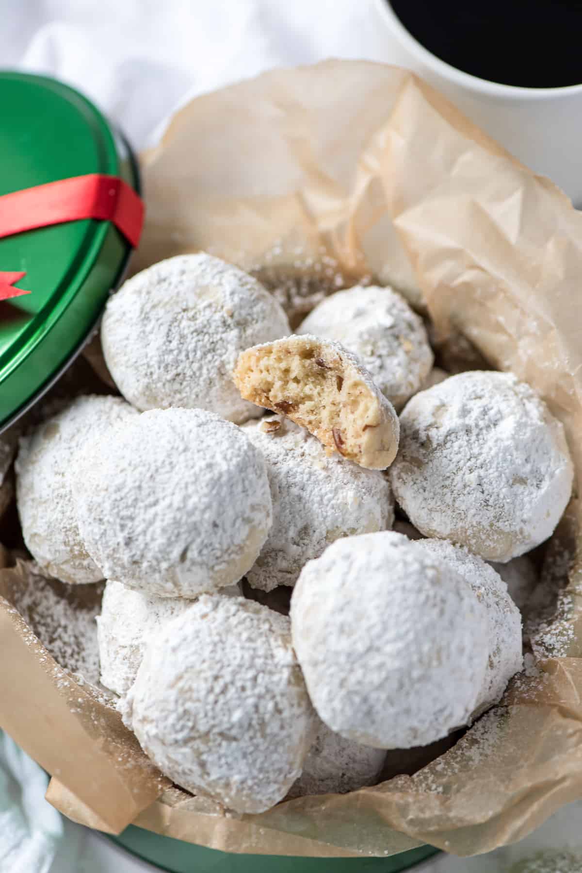 A pile of powdered sugar dusted Pecan Sandies in a green cookie tin with a bite missing from the cookie on top.