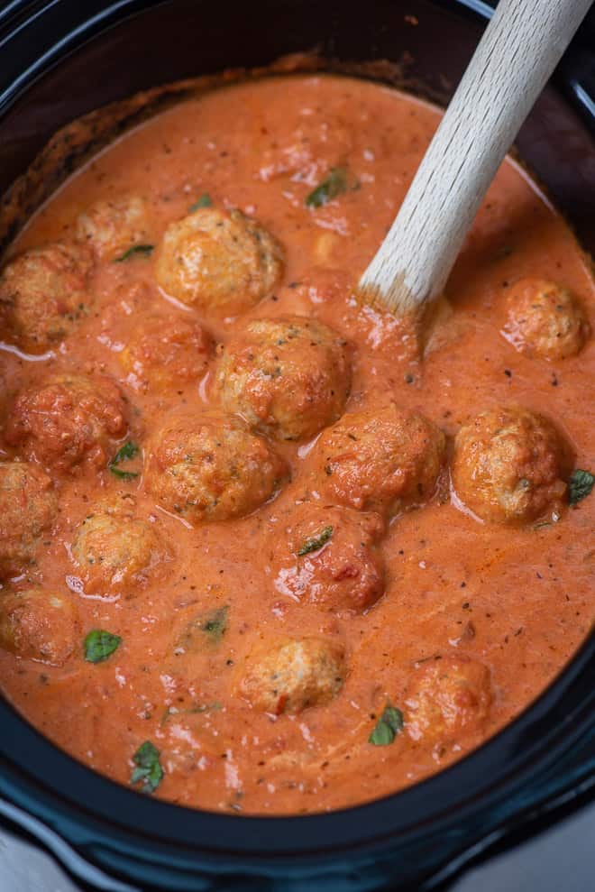 A slow cooker filled with chicken meatballs being stirred with a wooden spoon.