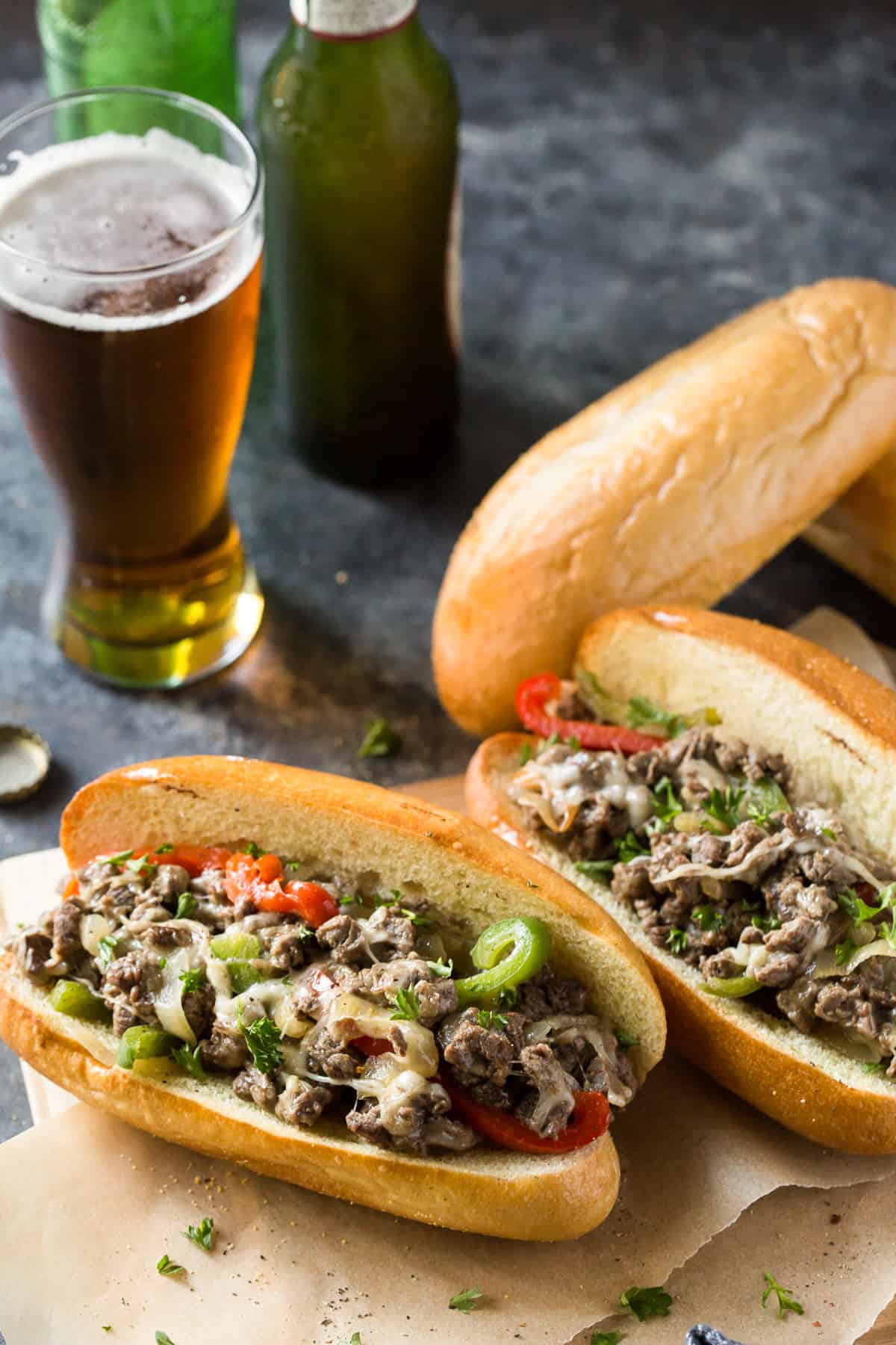 Two Philly Cheesesteak Sandwiches with peppers and onions with a glass of beer in the background.