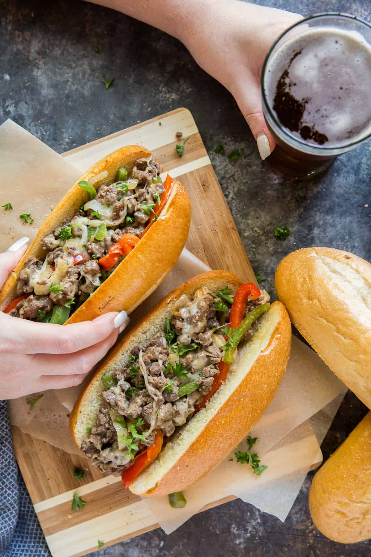 A hand holding a Philly cheesesteak while the other hand holds a glass of beer.