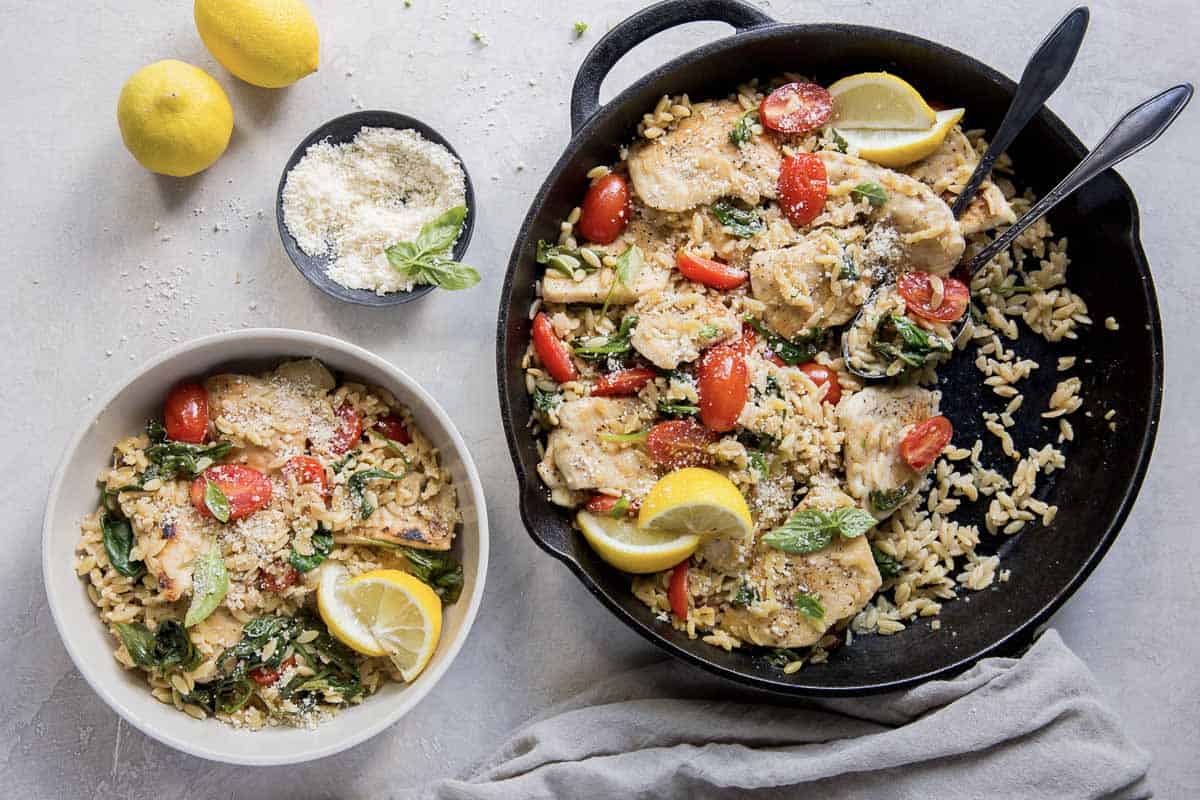 A skillet and a bowl are filled with a chicken and rice dish. The meal is garnished with lemon slices, cherry tomatoes, and fresh herbs. A small bowl containing grated cheese and two whole lemons are placed beside the plates on a light gray surface.