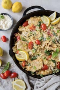 A skillet filled with orzo, chicken, grape tomatoes, and slices of lemon.