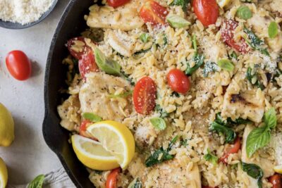 A skillet filled with orzo, chicken, grape tomatoes, and slices of lemon.