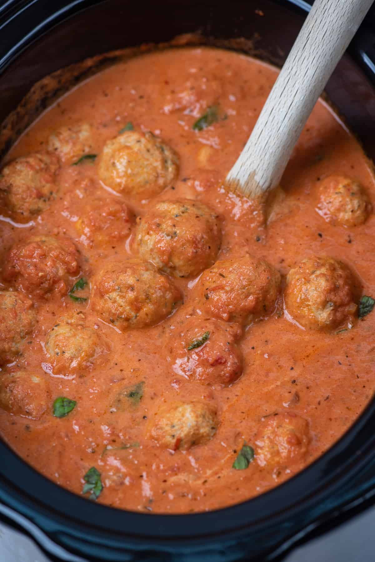 A wooden spoon resting in a slow cooker filled with chicken meatballs in tomato cream sauce.