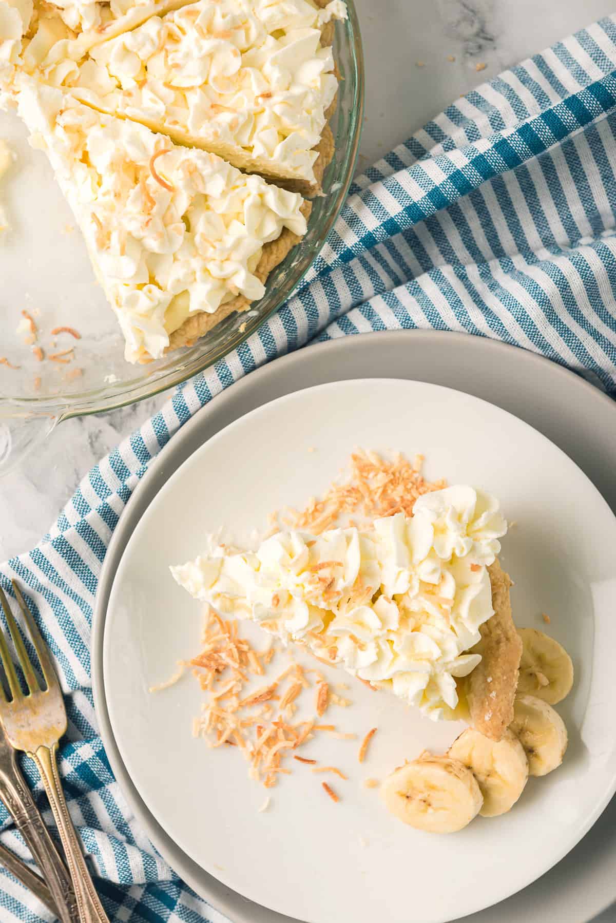 An overhead shot of a slice of banana cream pie on a plate on top of a blue striped kitchen towel.