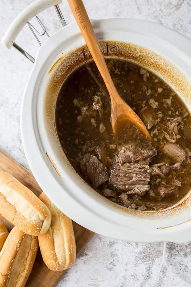 The beef is shredded into the cooking liquid in a white slow cooker.