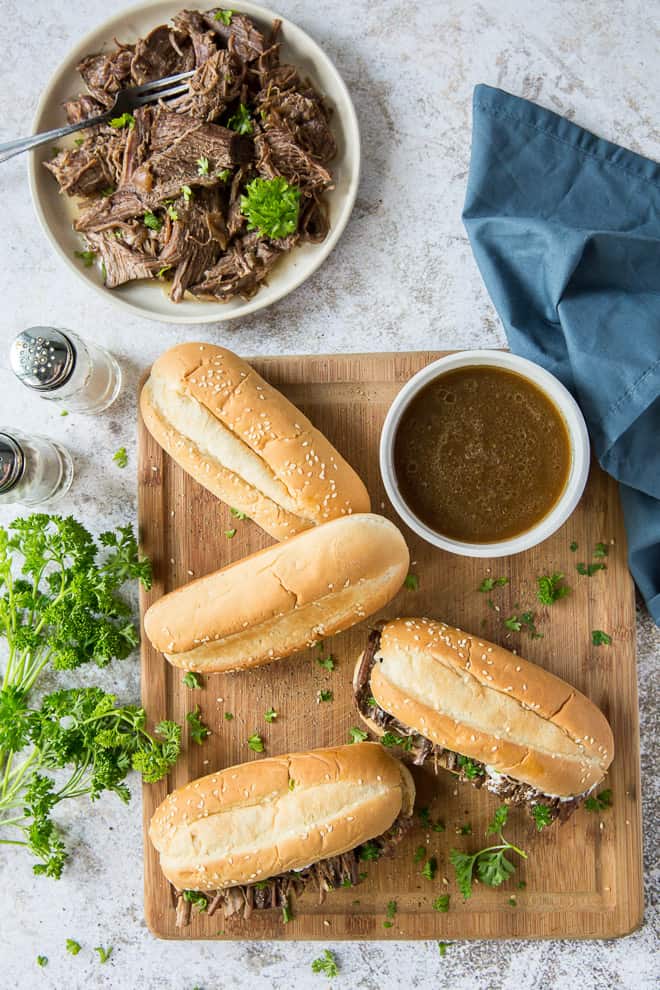 An overhead shot of a cutting board full of sandwiches, a bowl of au jus, and a bowl full of shredded beef.