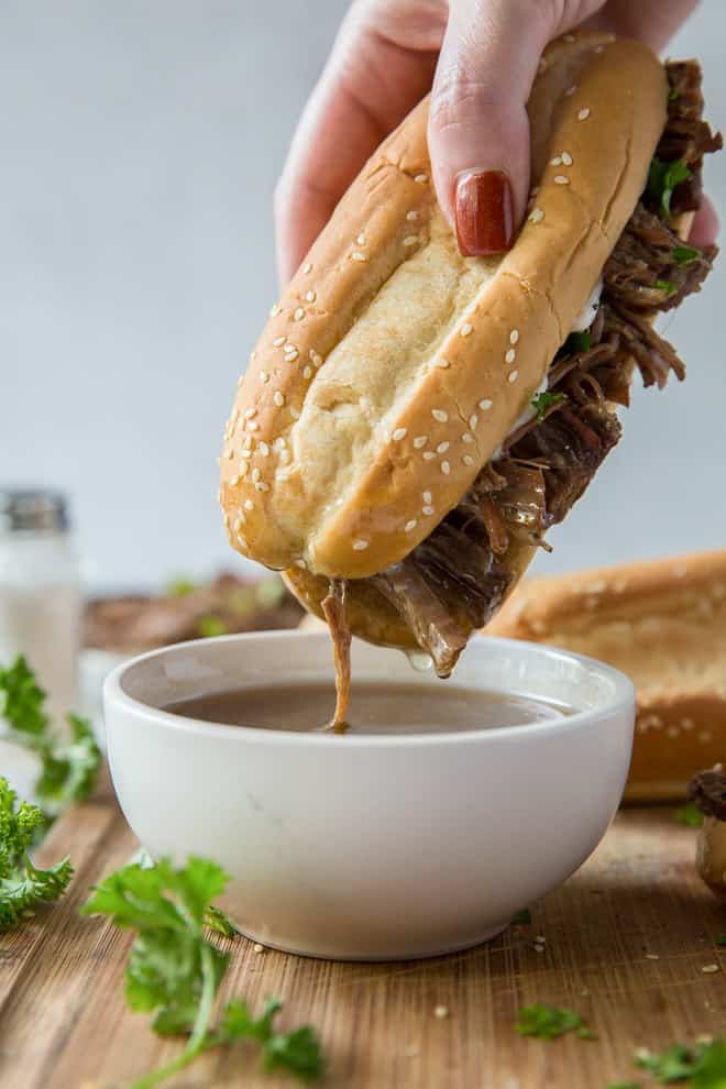 A slow cooker French Dip Sandwich is dipped into a bowl of au jus.