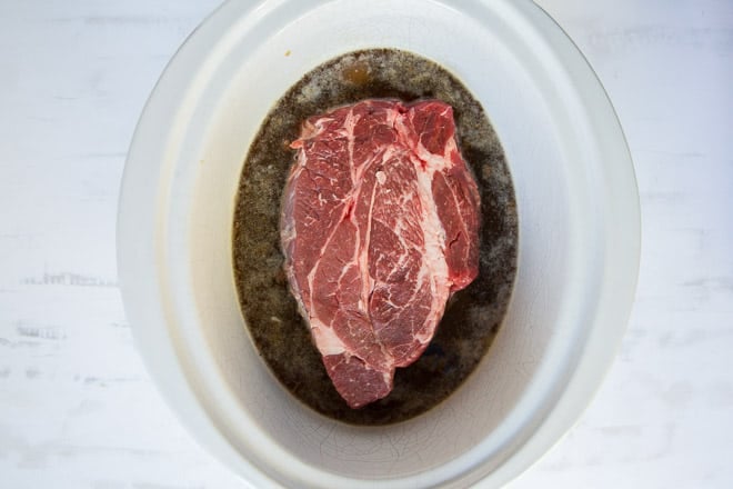 An overhead shot of a chuck roast in cooking liquid in a white slow cooker insert.