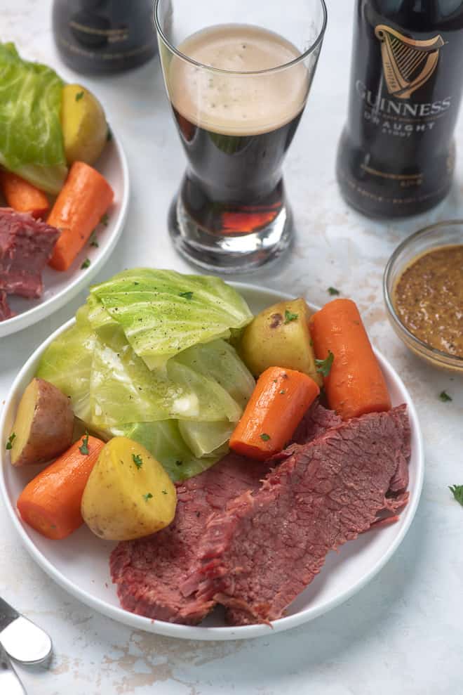 A serving of Instant Pot Corned Beef and Cabbage with carrots and potatoes on a white serving dish with a glass full of Guinness in the background.