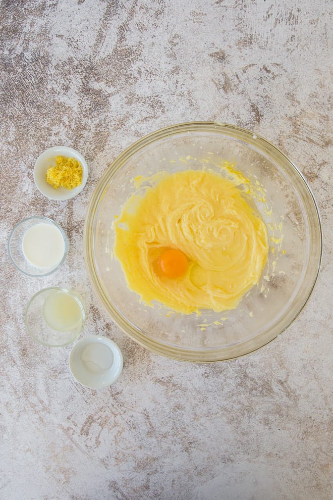 An egg is dropped into the wet mixture in a large glass mixing bowl with smaller bowls full of ingredients next to it.
