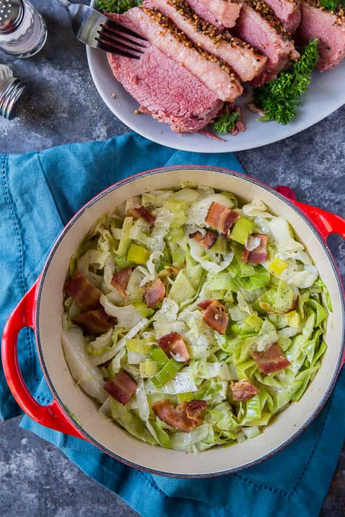 A red Dutch oven filled with Sauteed Cabbage with Leeks and Bacon shot from over the top with a platter of corned beef behind it.