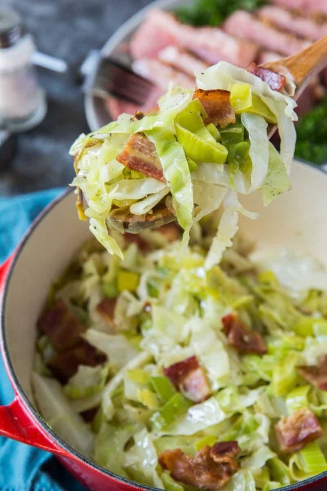 A wooden spoon lifts up some of the sauteed cabbage from a red Dutch oven.