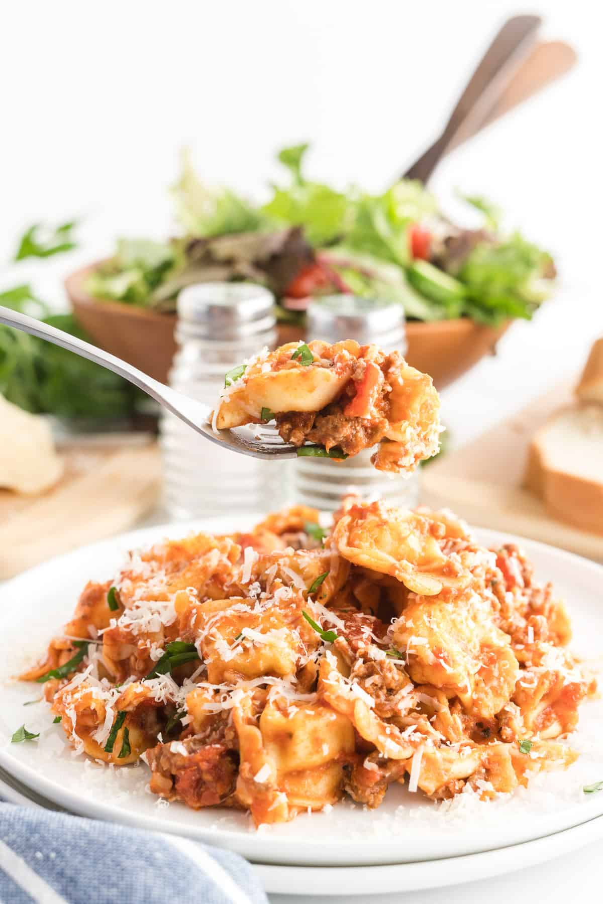 A fork lifts a spoonful of tortellini from a white plate with a green salad in the background.