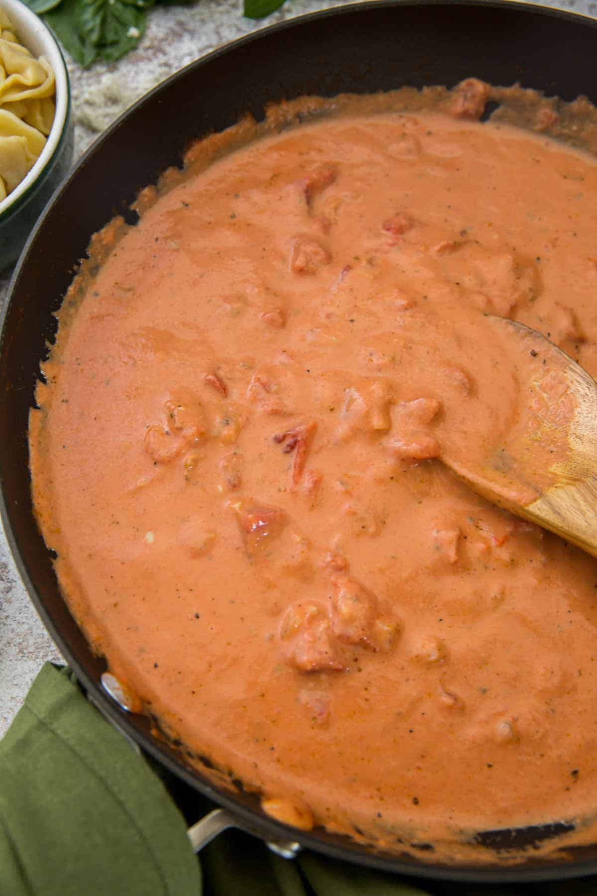 A woodens poon resting in a skillet filled with tomato cream sauce.