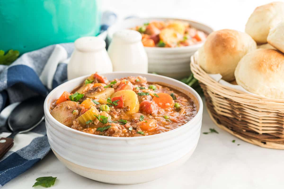 Hamburger Stew with Rice in a white serving bowl with a basket of rolls in the background.