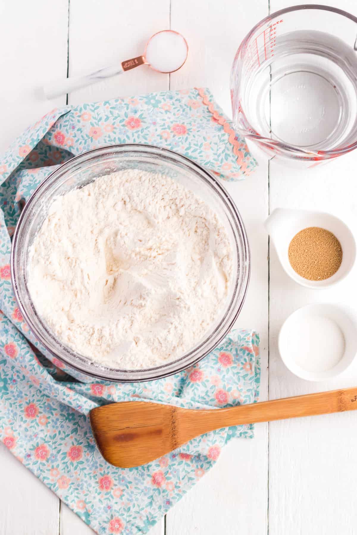 The ingredients for Homemade English Muffins on a white board with a blue kitchen towel and wooden spatula.