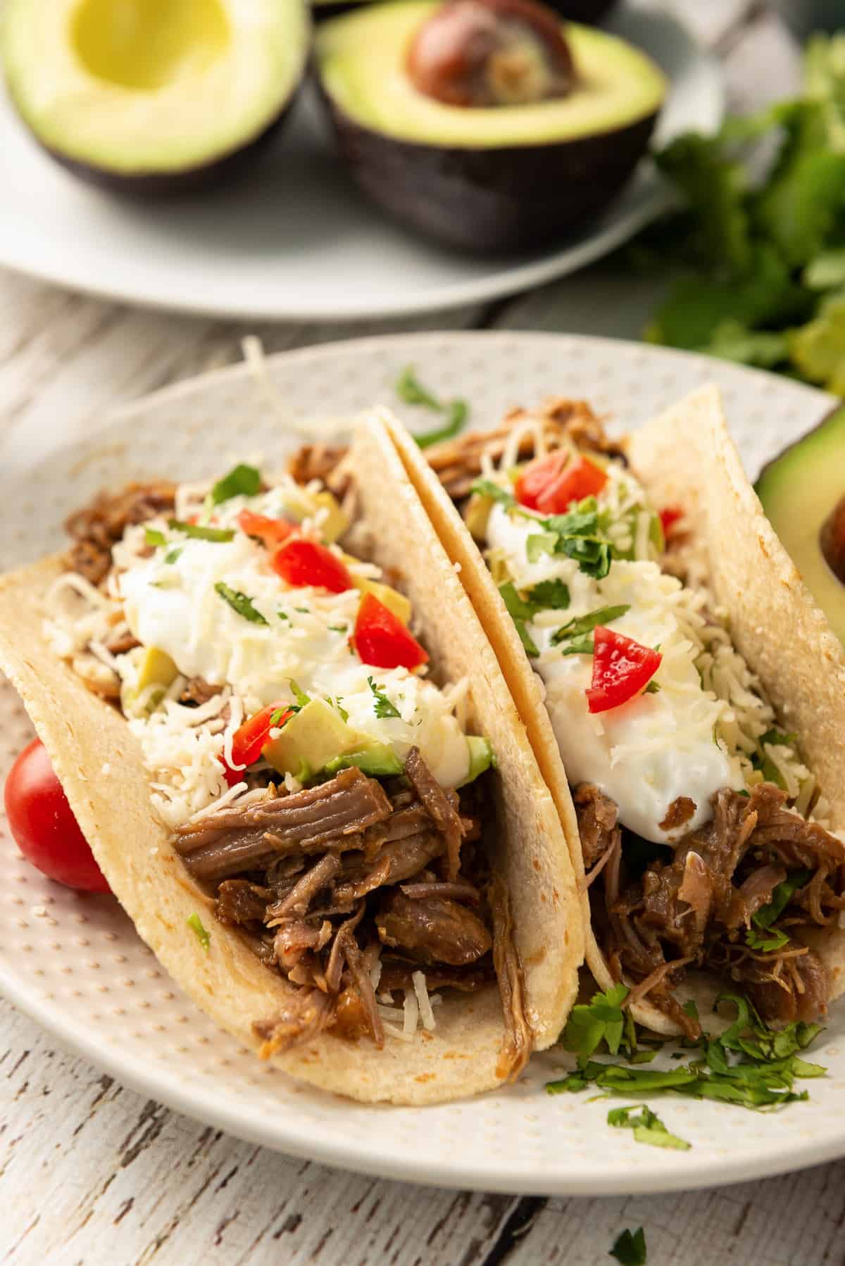 A white serving plate with two Chipotle Pork Street Tacos with halved avocados in the background.