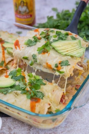 A spatula lifts enchiladas from the dish with hot sauce in the background.