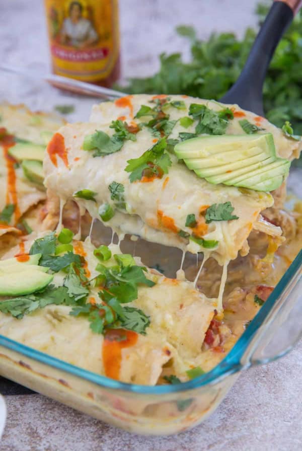 A spatula lifts enchiladas from the dish with hot sauce in the background.