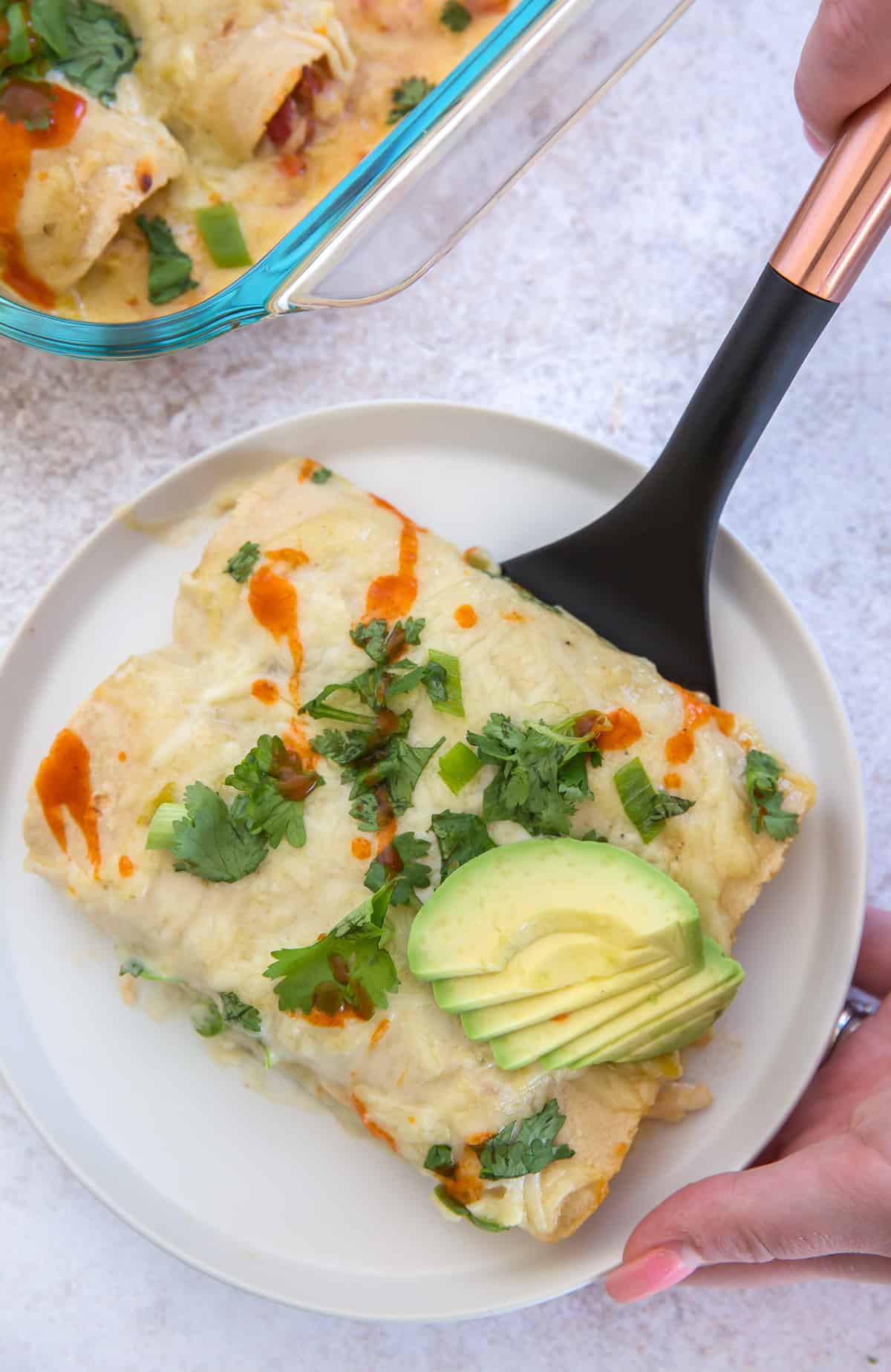 A hand holding a spatula places two enchiladas on a white serving plate.