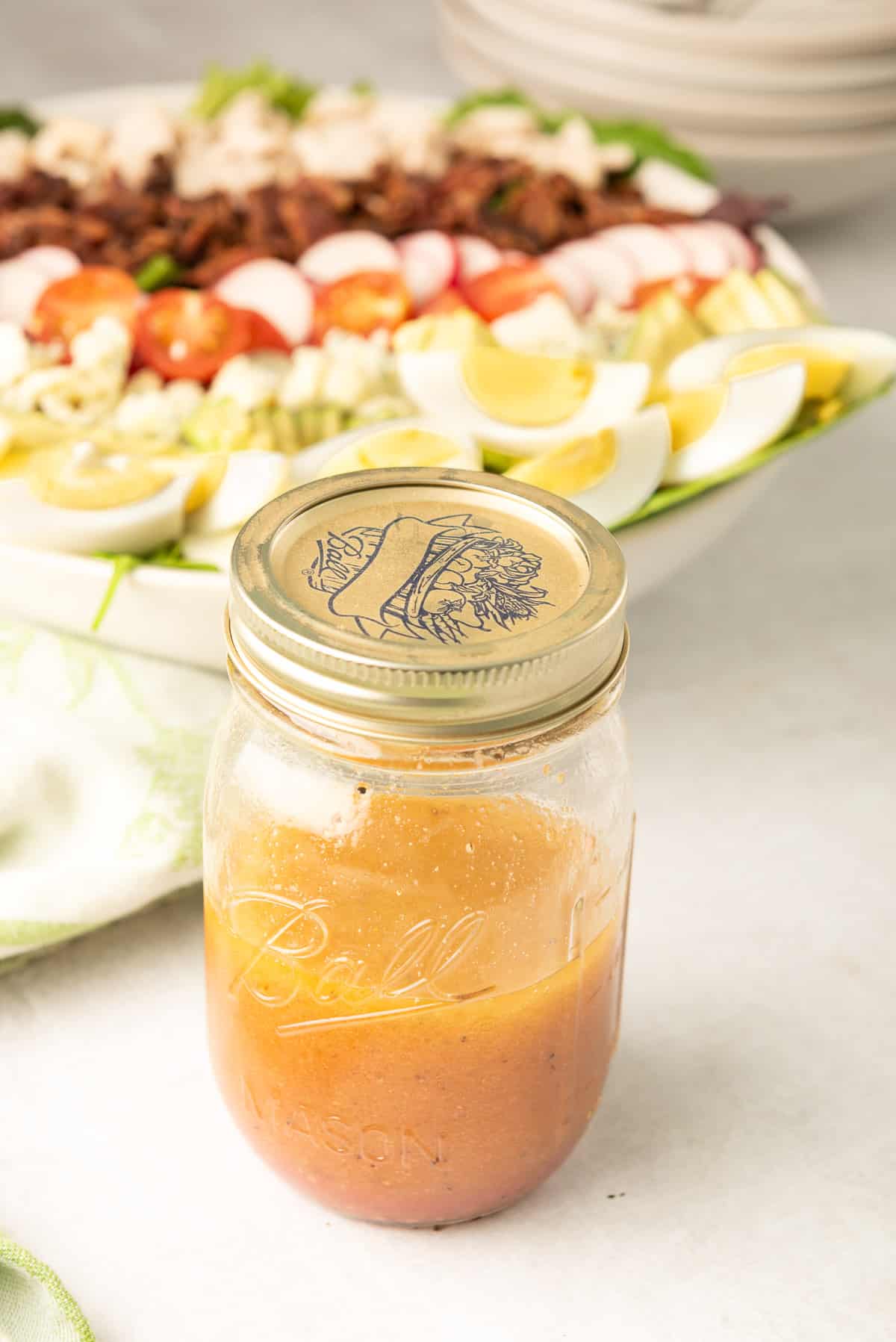 A mason jar filled with the French Vinaigrette with a Chicken Cobb Salad in the background.