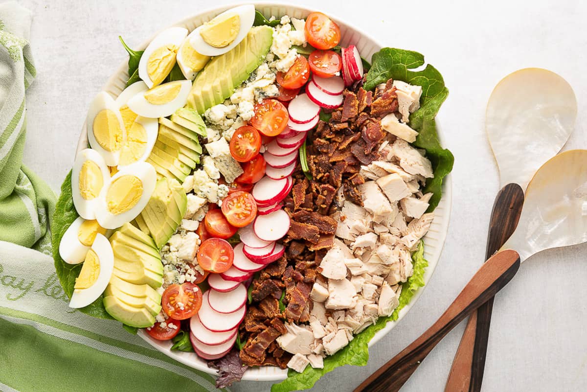 An over the top shot of the Easy Cobb Salad ingredients layered in a large white serving bowl with utensils laying next to it.