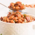 A spoon lifts some baked beans from a white serving dish.