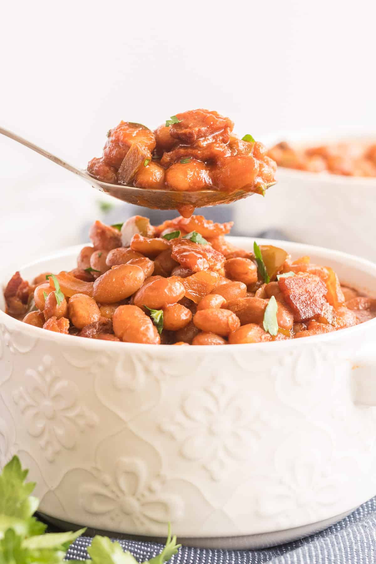 A spoon lifts some baked beans from a white serving dish.