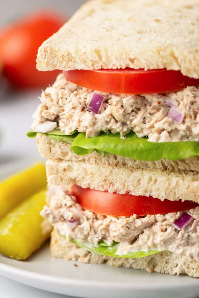 A close up of a Lemon Pepper Tuna Sandwich layered with tomatoes and lettuce on a white plate with pickles.