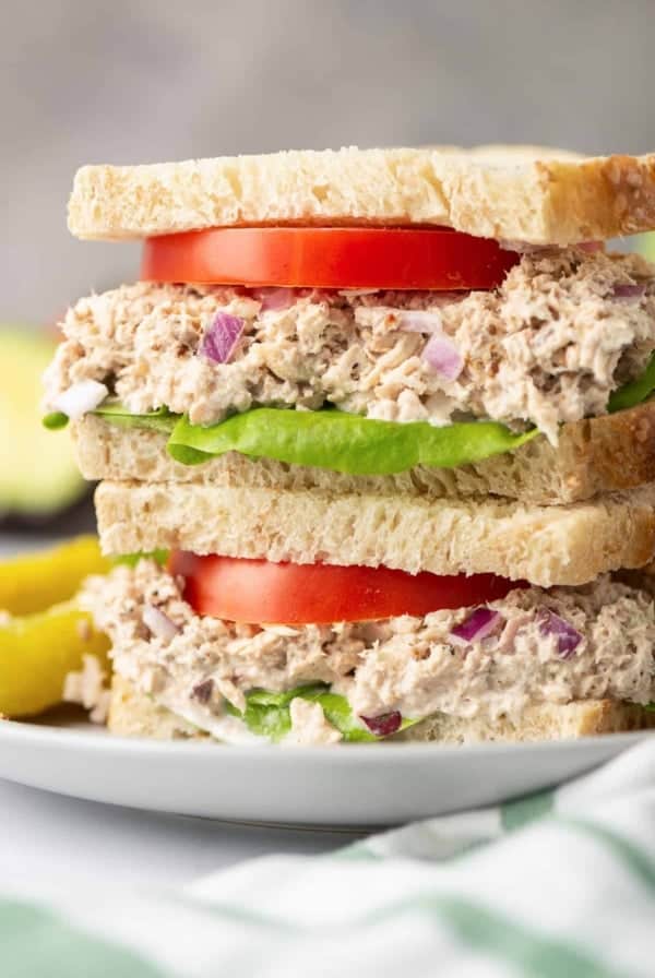 A Lemon Pepper Tuna Sandwich cut in half and stacked on a white plate with pickles.