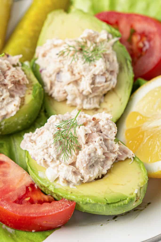 Avocado halves stuffed with Lemon Pepper Tuna on a plate with slices of tomato and lemon.