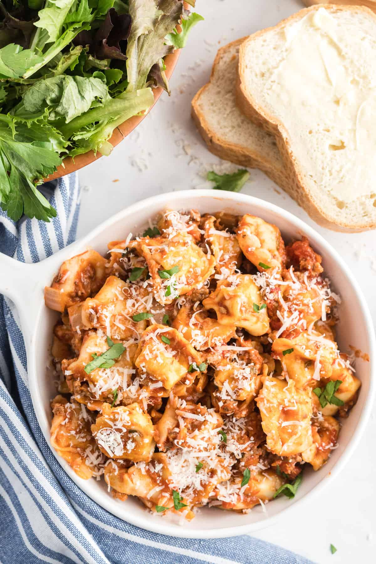 A small white handled bowl full of tortellini with buttered bread and a green salad in the background.