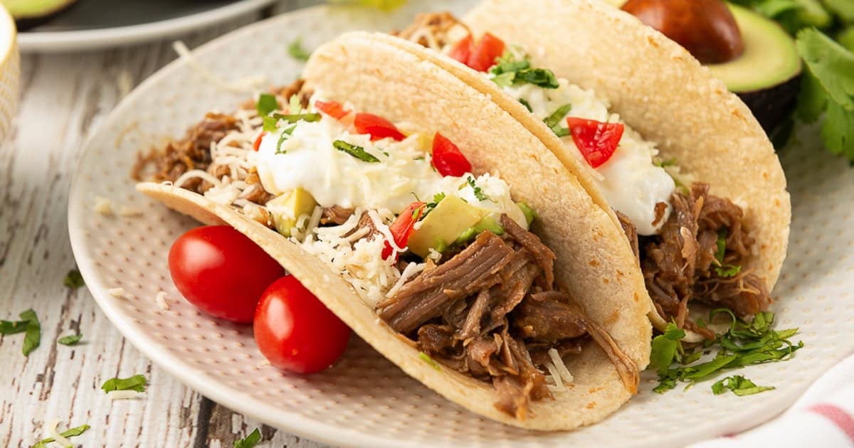 Shredded pork tacos with cheese, tomatoes, and sour cream on a white plate.