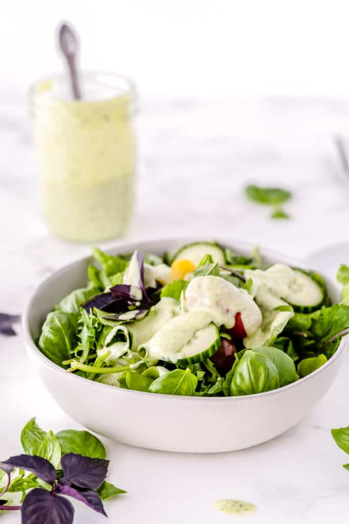 A white bowl filled with green salad with a mason jar full of dressing behind it.