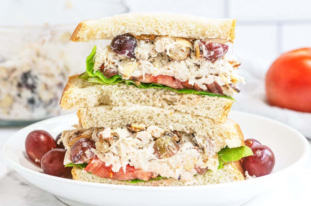 A Napa Almond Chicken Salad Sandwich sliced in half and stacked on a plate with a tomato in the background.
