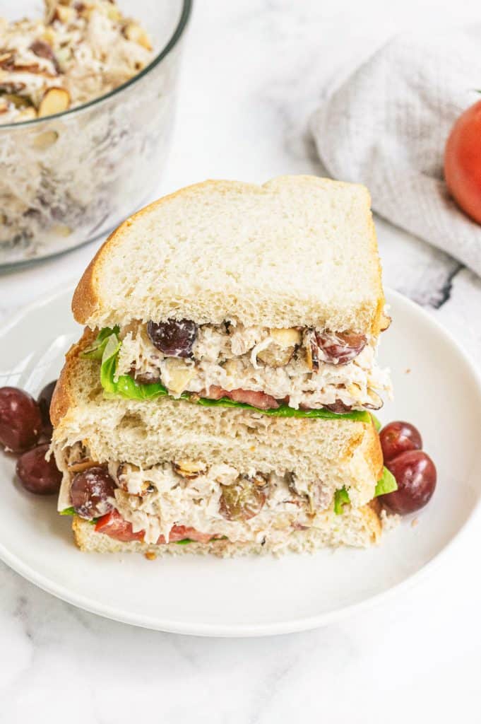 A Panera copycat chicken salad sandwich sliced in half and stacked on a white plate with grapes.