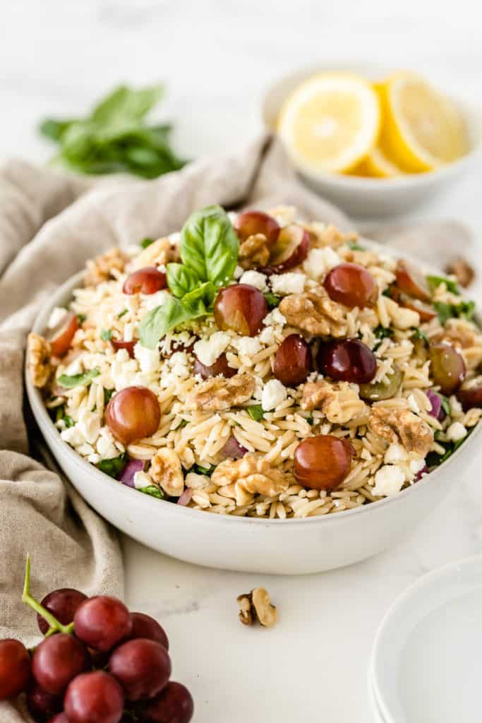 A serving bowl filled with the orzo pasta salad with lemons and grapes set around it.