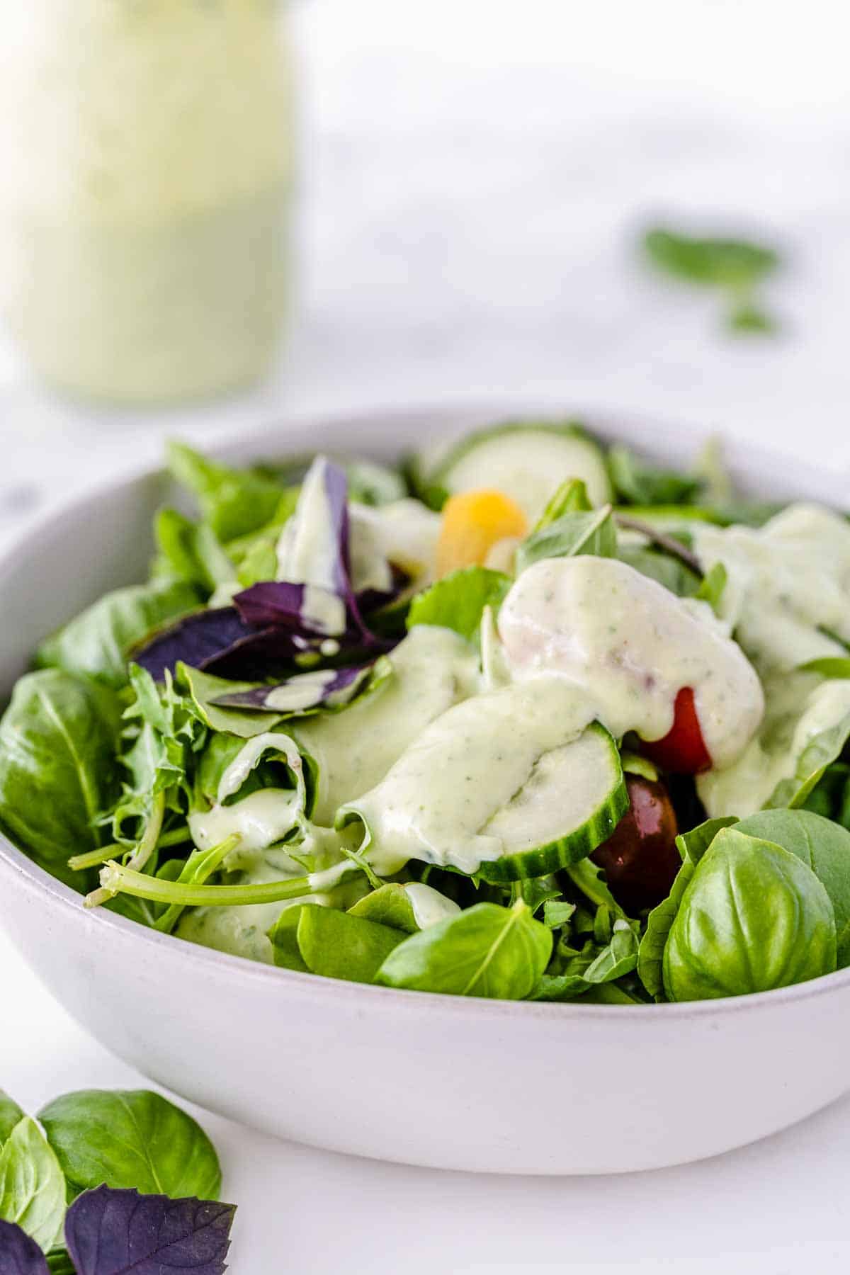 A white bowl filled with green salad with a mason jar full of dressing behind it.