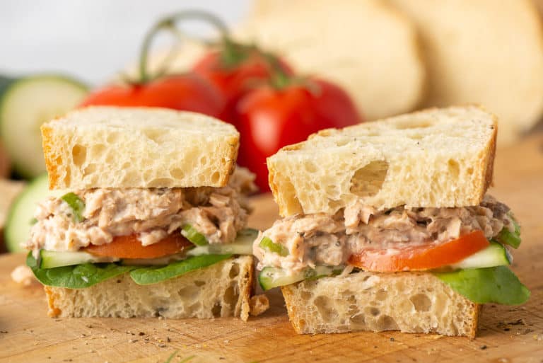 A sandwich with chicken salad, lettuce, tomato, and cucumber on rustic bread, halved and placed on a wooden surface. Blurred tomatoes and cucumber in the background.