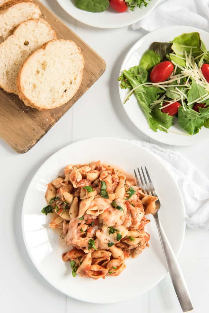 Pasta on a white plate with a salad and bread in the background.