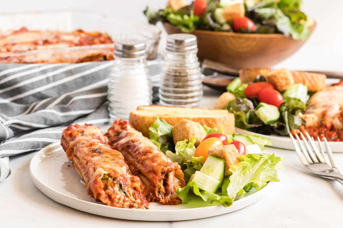 A plate of manicotti and salad in front on a dinner table.